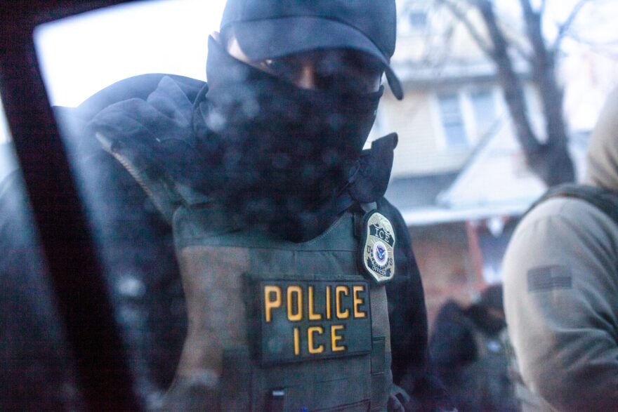 Masked ICE officer in tactical gear photographed through a vehicle window